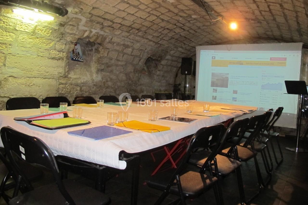 Salle voûtée avec une grande table rectangulaire dressée pour une réunion, équipée d'un projecteur et de chaises noires.