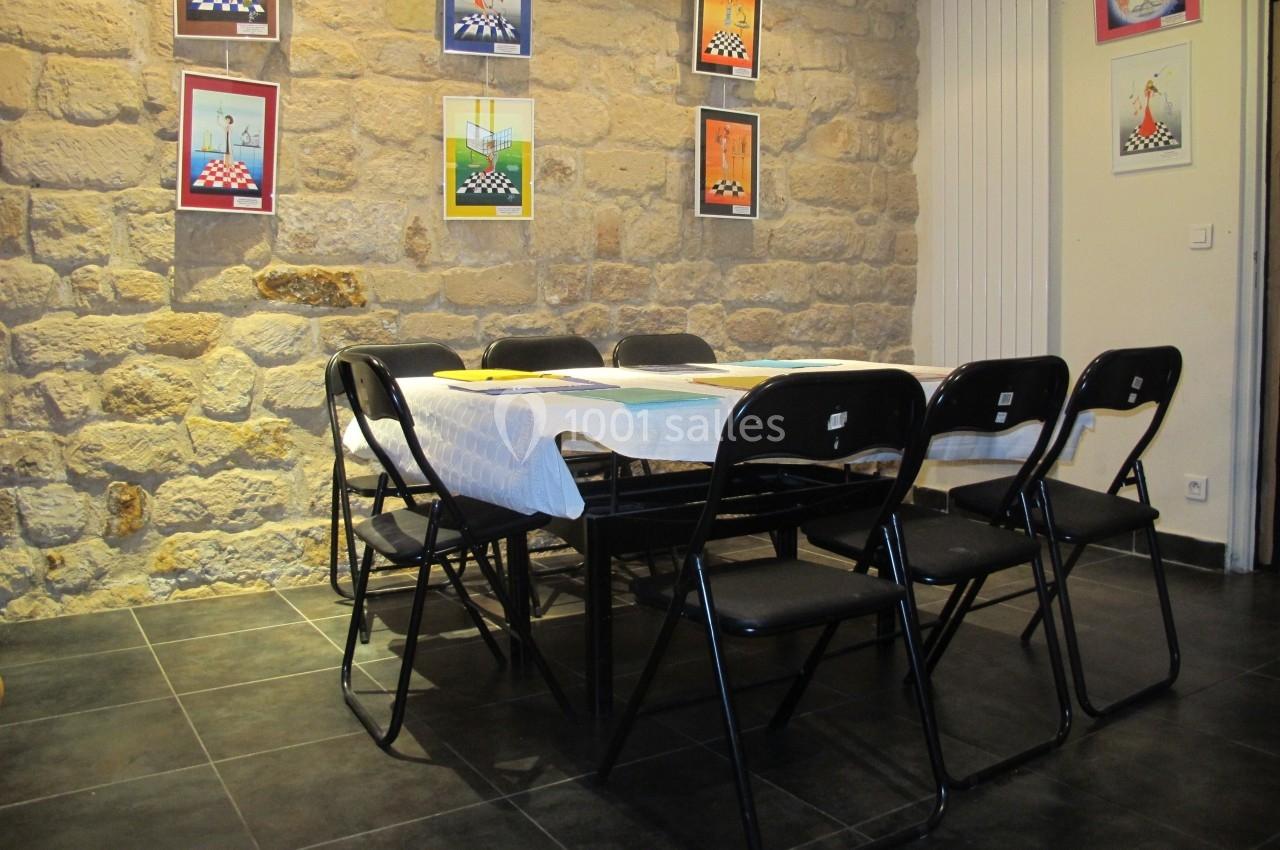 Salle avec murs en pierre, table couverte d'une nappe blanche entourée de chaises noires pliantes, tableaux accrochés au mur.