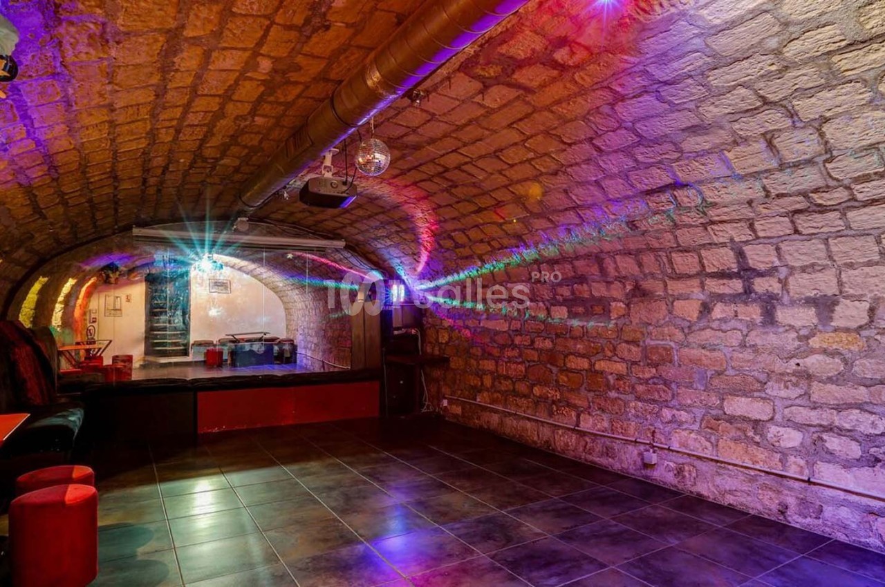 Salle voûtée en pierre avec éclairage coloré, petite scène et mobilier minimaliste.