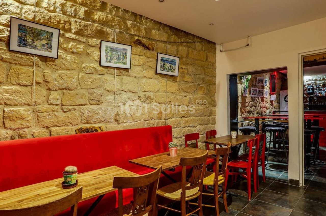 Salle de restaurant avec murs en pierre, banquette rouge, tables en bois et vue sur un bar éclairé en rouge.