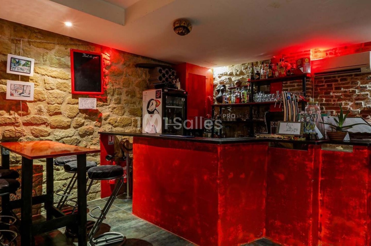 Bar avec comptoir rouge éclairé, tabourets hauts, murs en pierre et briques, décorations murales et étagères avec bouteilles.