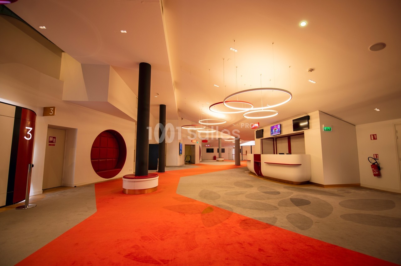 Couloir lumineux avec moquette orange, comptoirs d'accueil, panneaux numérotés et éclairage circulaire suspendu.