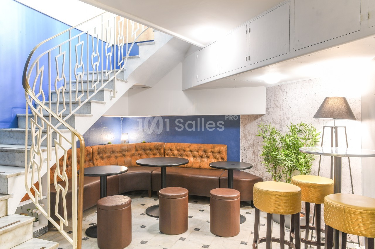 Escalier en colimaçon menant à un espace lounge avec banquette en cuir, tables rondes et tabourets dans un décor lumineux.
