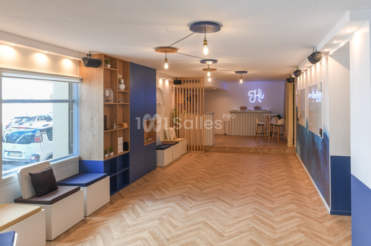 Salle lumineuse avec parquet en chevrons, banquettes près d'une fenêtre, et comptoir avec enseigne lumineuse au fond.