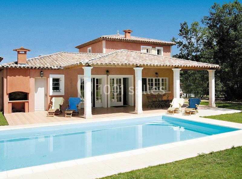 Maison méditerranéenne avec piscine, terrasse aménagée et chaises longues, entourée de verdure sous un ciel bleu.