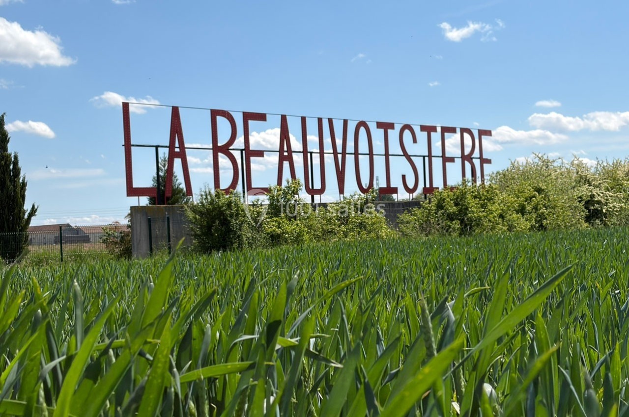 Panneau rouge ’La Beauvoisière’ sur une structure métallique, entouré de végétation et d'un champ sous un ciel bleu.
