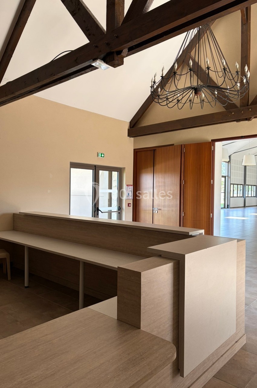 Accueil en bois clair avec comptoirs, situé dans une salle lumineuse aux poutres apparentes et lustre suspendu.