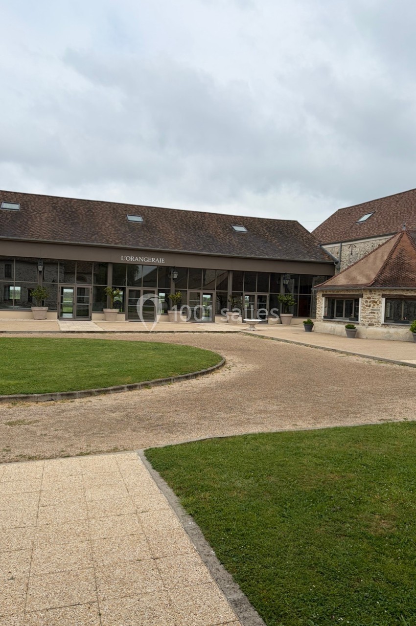 Bâtiment en pierre avec toit en tuiles, grandes baies vitrées et allée pavée entourée de pelouse.