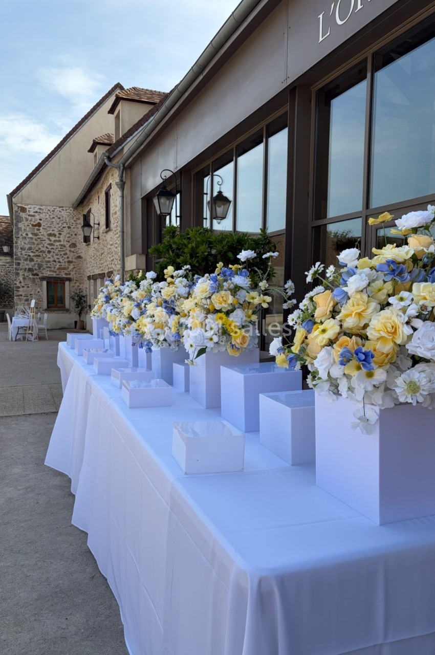 Table décorée de bouquets de fleurs jaunes, blanches et bleues devant un bâtiment en pierre sous un ciel dégagé.
