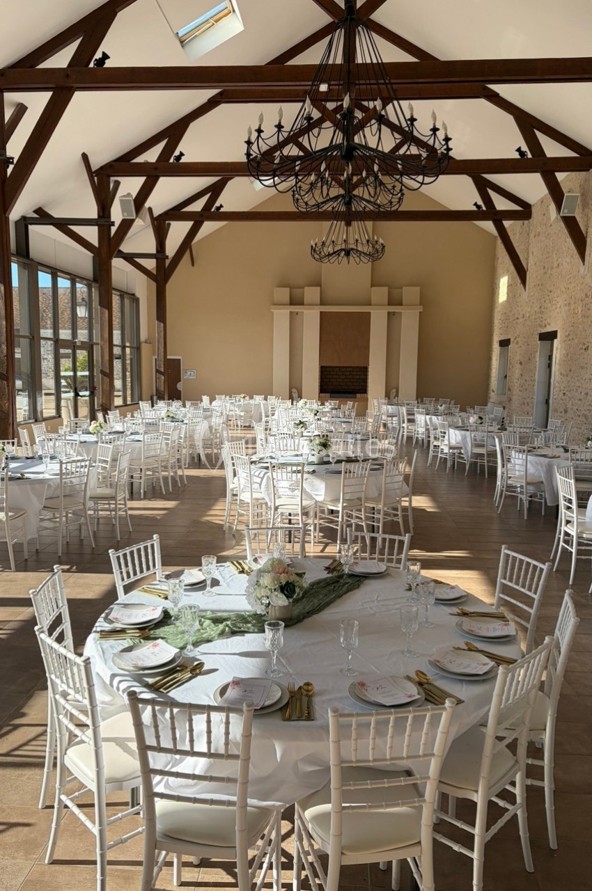 Salle de réception lumineuse avec tables rondes dressées, chaises blanches et poutres apparentes en bois.
