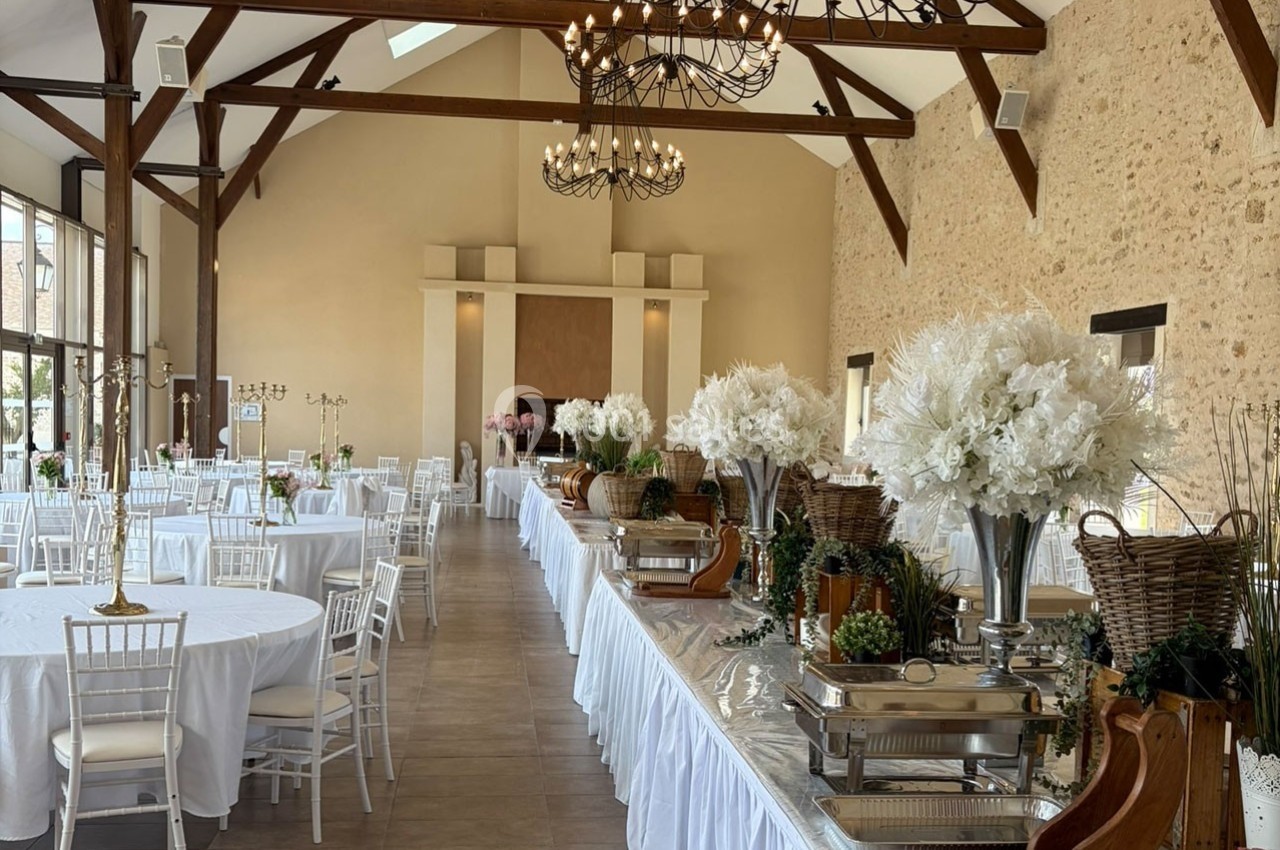 Salle de réception lumineuse avec tables rondes dressées, décorations florales blanches et buffet disposé sur le côté.