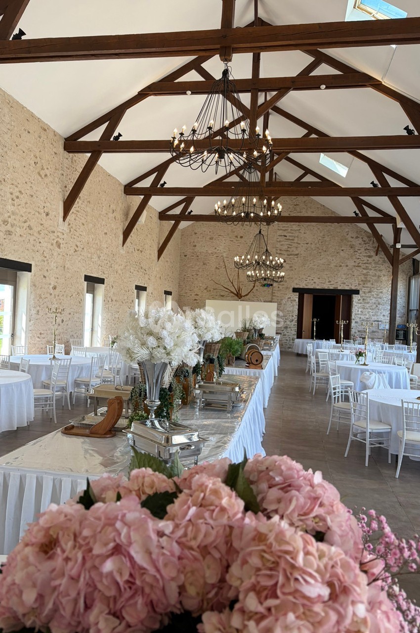 Salle de réception lumineuse avec poutres apparentes, tables décorées de fleurs blanches et chaises blanches.