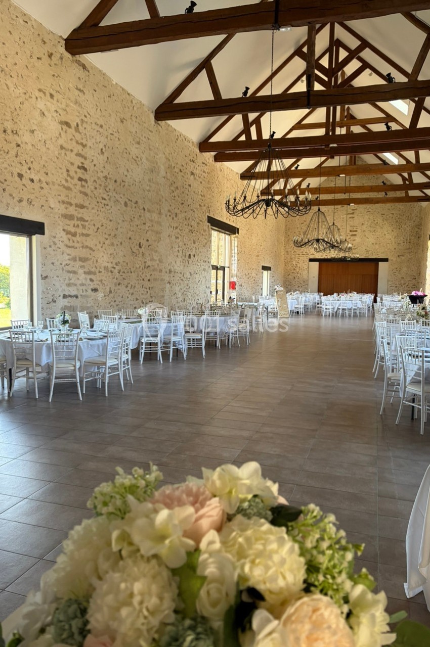 Salle de réception spacieuse avec murs en pierre, poutres apparentes, tables dressées et bouquet de fleurs au premier plan.
