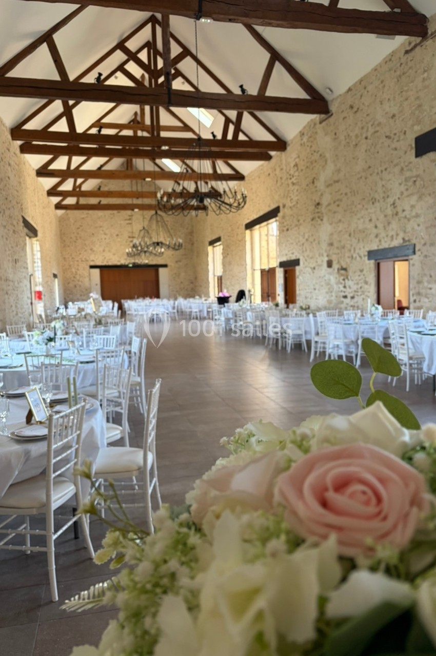 Grande salle de réception en pierre avec tables décorées de nappes blanches et chaises, vue sur un bouquet au premier plan.