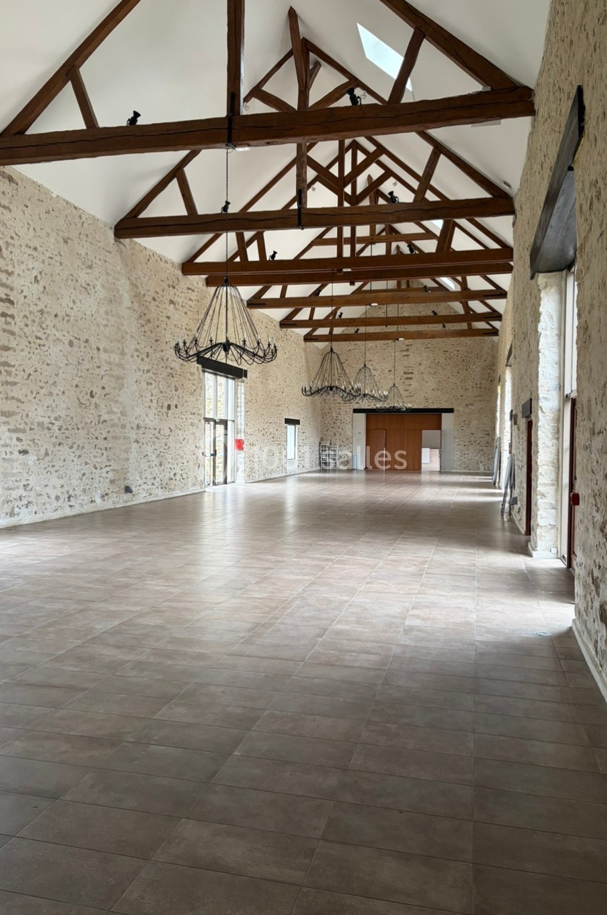 Grande salle avec murs en pierre apparente, poutres en bois au plafond et lustres suspendus, sol carrelé.