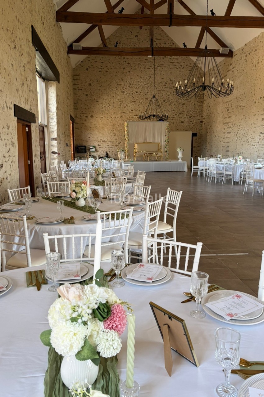 Salle de réception en pierre avec tables dressées, chaises blanches et décorations florales élégantes.