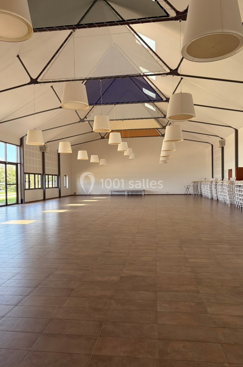 Grande salle vide avec sol en carrelage, plafond haut, luminaires suspendus et chaises empilées sur le côté.