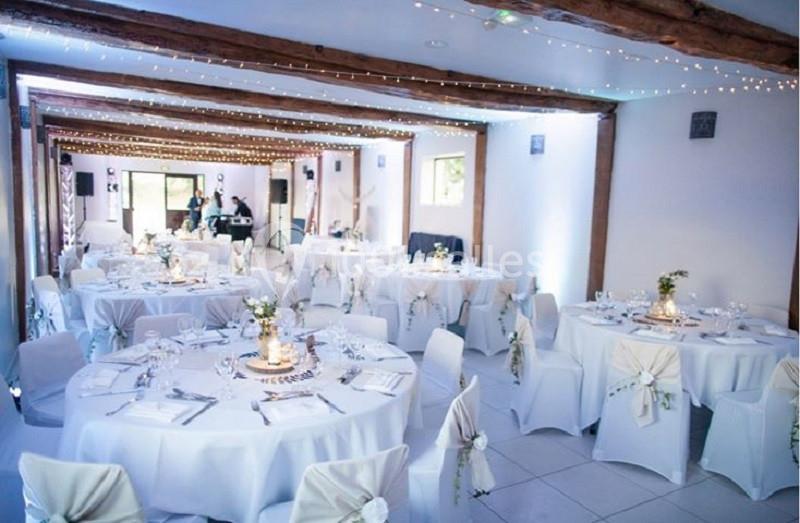 Salle de réception décorée avec des tables rondes, nappes blanches, chaises ornées et guirlandes lumineuses au plafond.