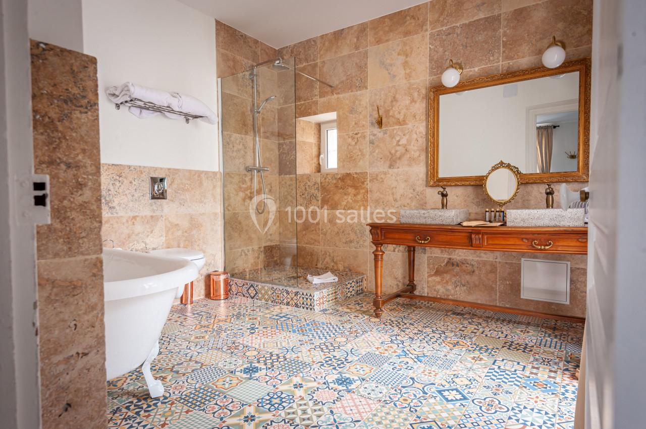 Salle de bain avec baignoire sur pieds, douche vitrée, carrelage coloré au sol et meuble en bois avec miroir doré.