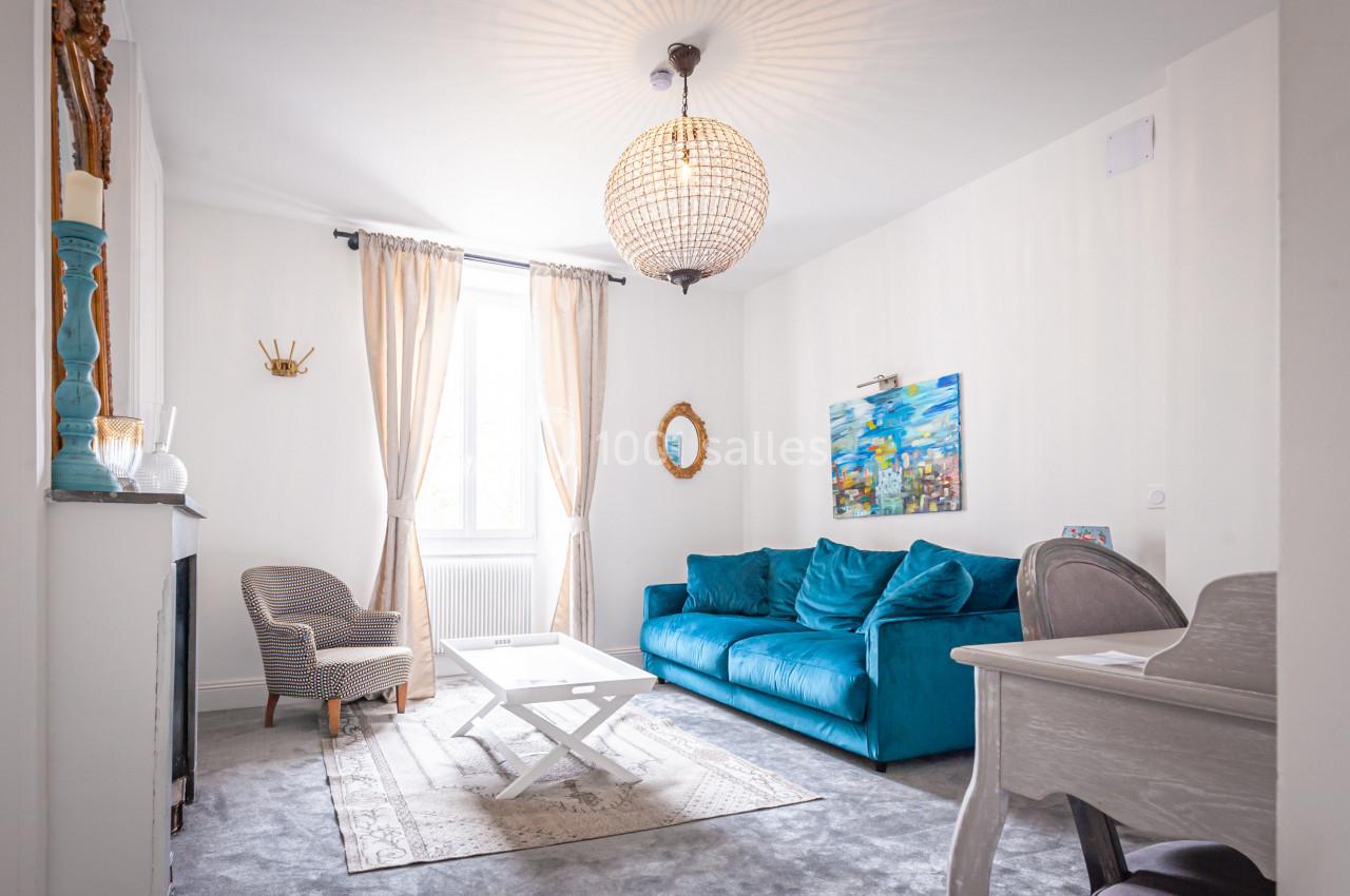 Salon lumineux avec canapé bleu, fauteuil rayé, bureau gris, table basse blanche et lustre suspendu.