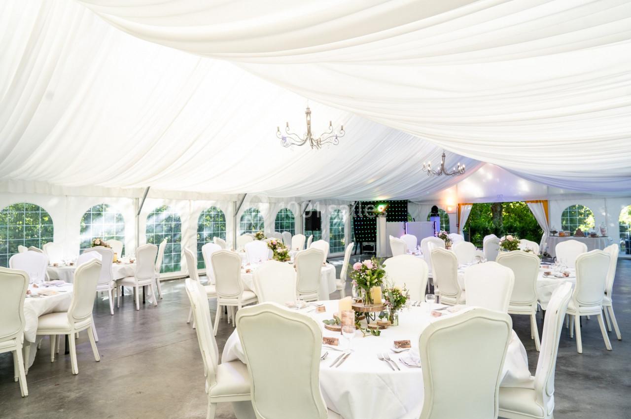 Salle de réception sous une tente blanche avec tables décorées, chaises blanches et lustres suspendus.