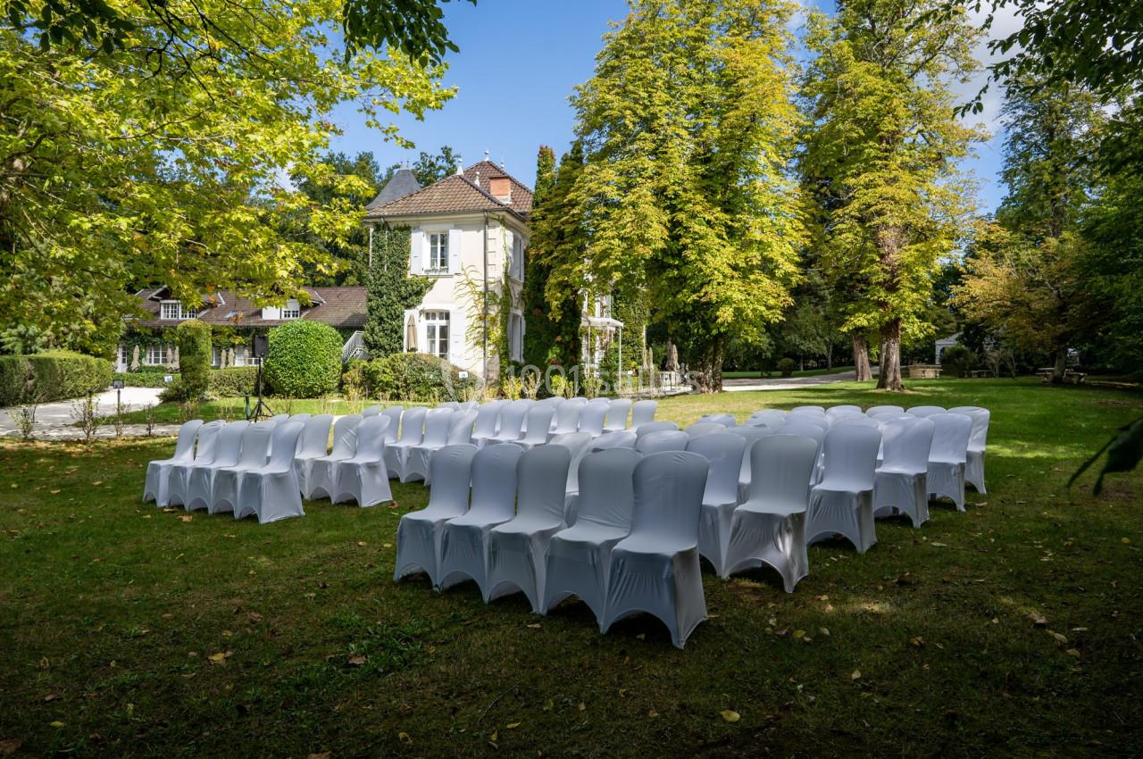 Chaises blanches disposées en cercle sur une pelouse, devant une maison entourée d'arbres en plein jour.