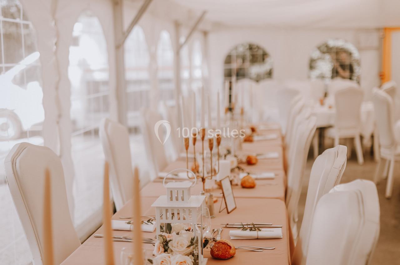 Table décorée pour un événement, avec nappes roses, bougies, fleurs et chaises blanches sous une tente lumineuse.
