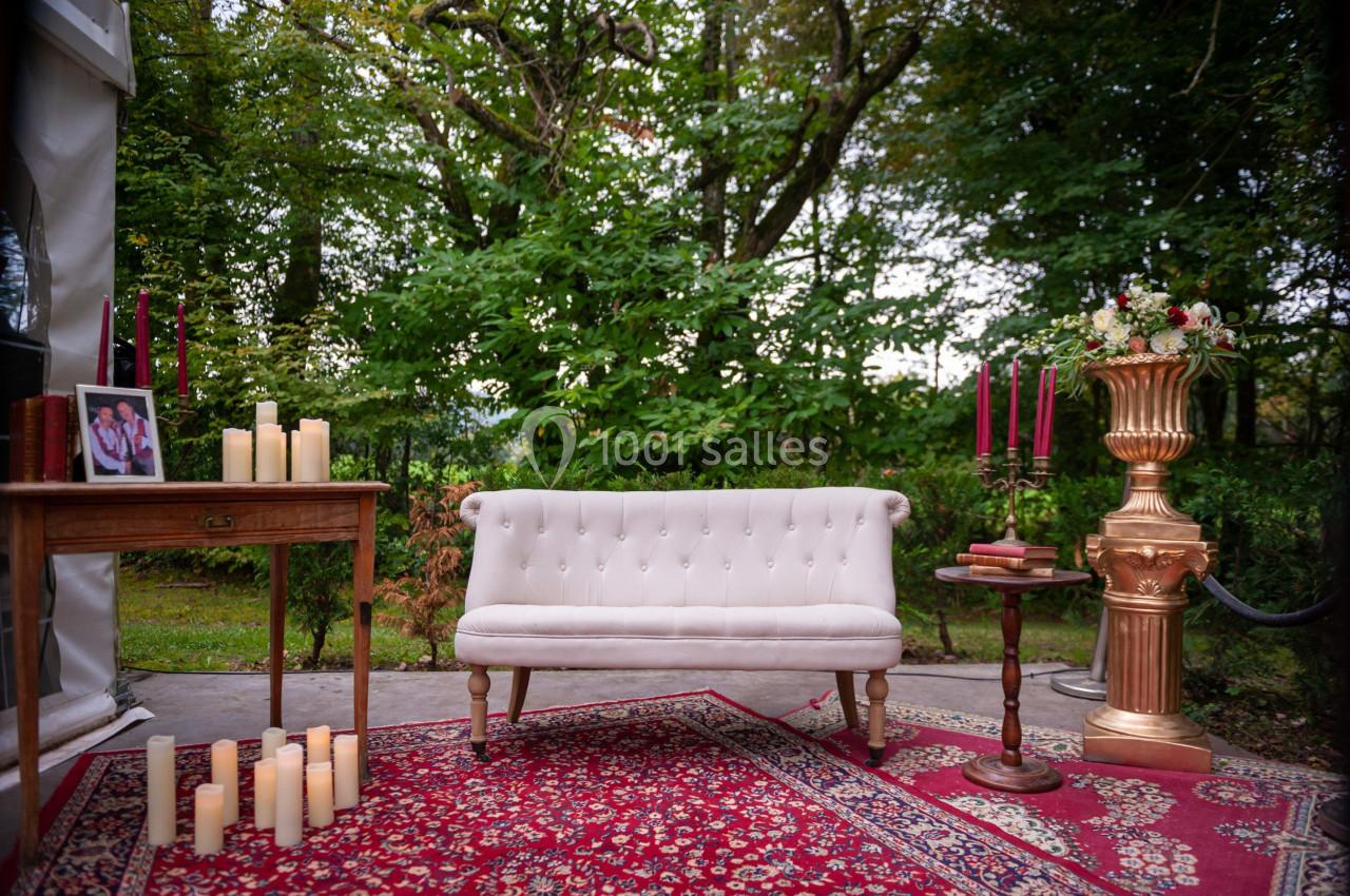 Canapé blanc capitonné sur un tapis rouge, entouré de bougies, d'une table en bois et d'un vase doré dans un jardin.