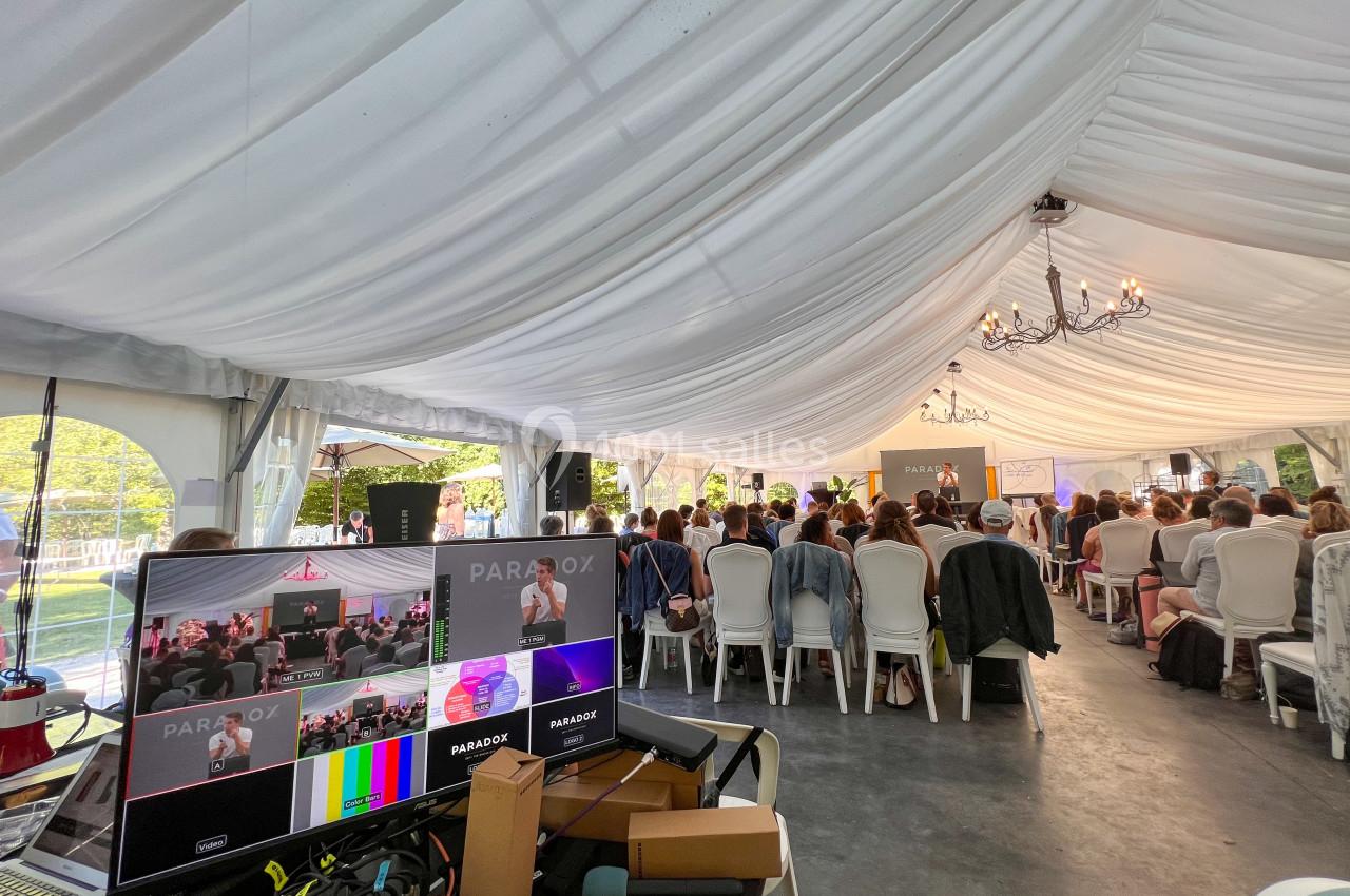 Vue d'une conférence sous une tente blanche, avec un public assis et un écran de contrôle au premier plan.