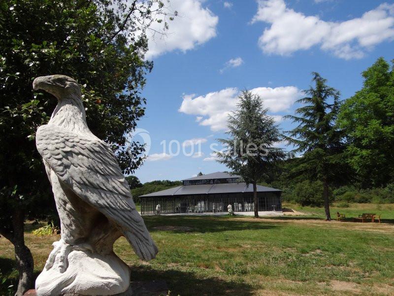 Statue d'aigle en pierre dans un parc verdoyant, avec une serre en arrière-plan sous un ciel partiellement nuageux.