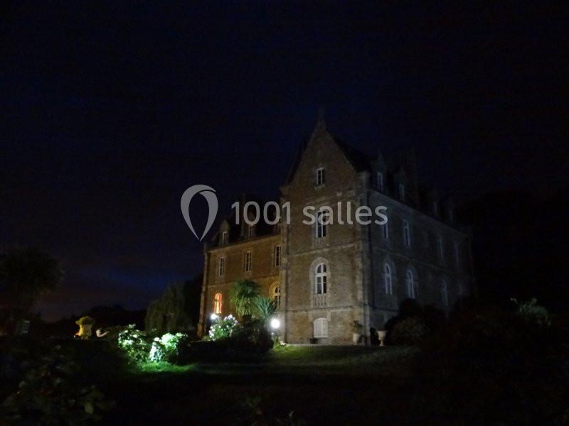 Manoir en pierre éclairé la nuit, entouré de végétation et d'un ciel sombre.