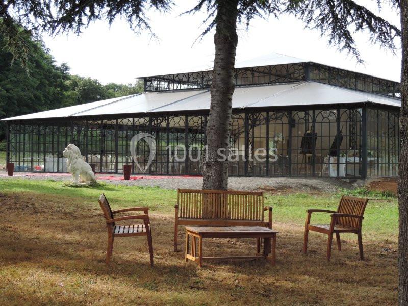 Structure en verre et métal avec un toit blanc, entourée d'arbres, avec des bancs en bois au premier plan.