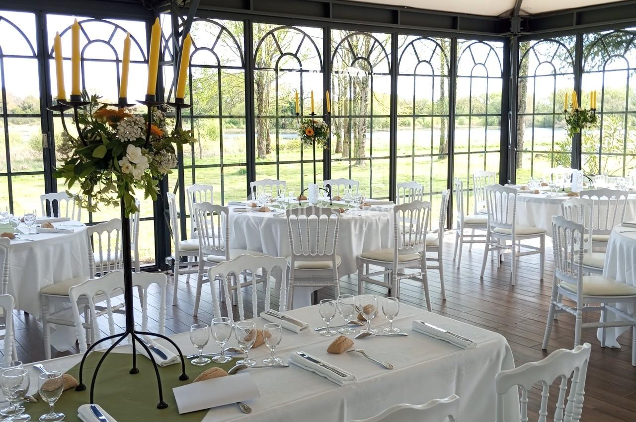 Salle lumineuse avec tables dressées, nappes blanches, chaises en bois, chandeliers et vue sur un jardin verdoyant.