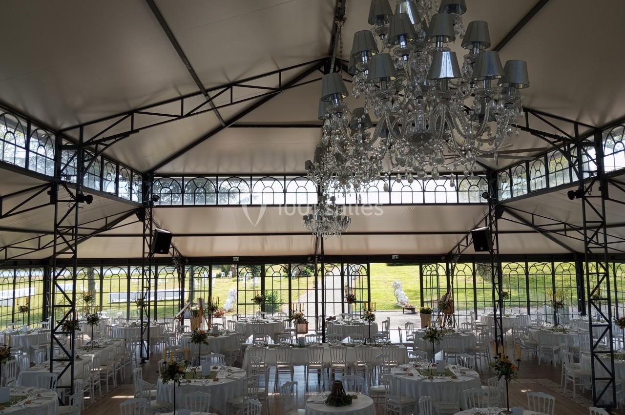 Salle de réception lumineuse avec tables rondes dressées, chaises blanches et lustres suspendus sous une verrière.