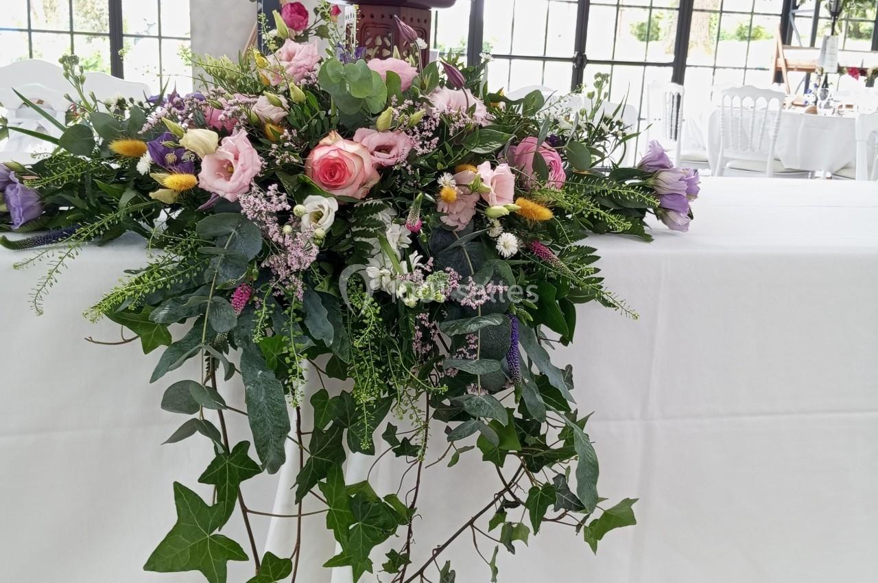 Arrangement floral coloré avec roses, feuillage et lierre, disposé sur une table blanche dans une salle lumineuse.