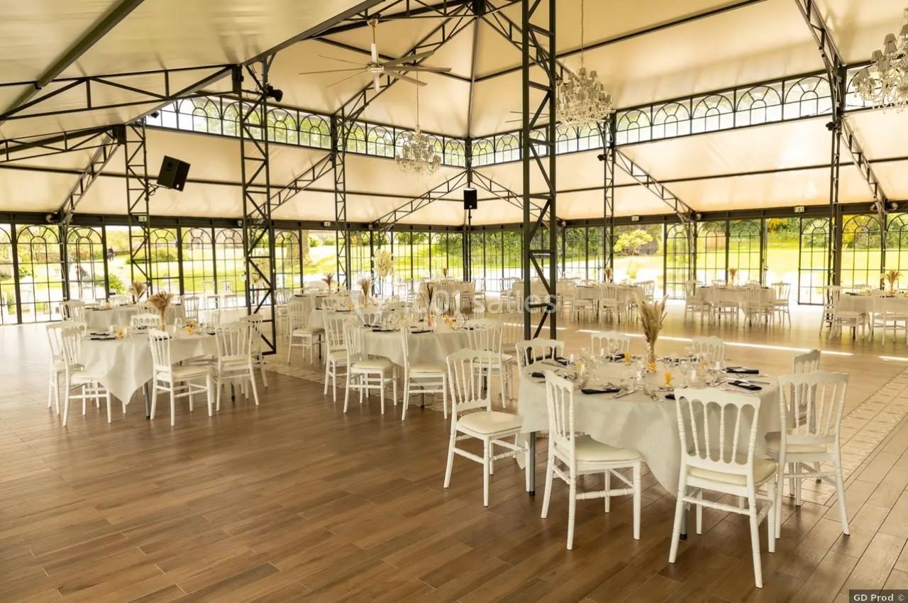 Salle de réception lumineuse avec tables rondes dressées, chaises blanches et structure métallique sous une verrière.