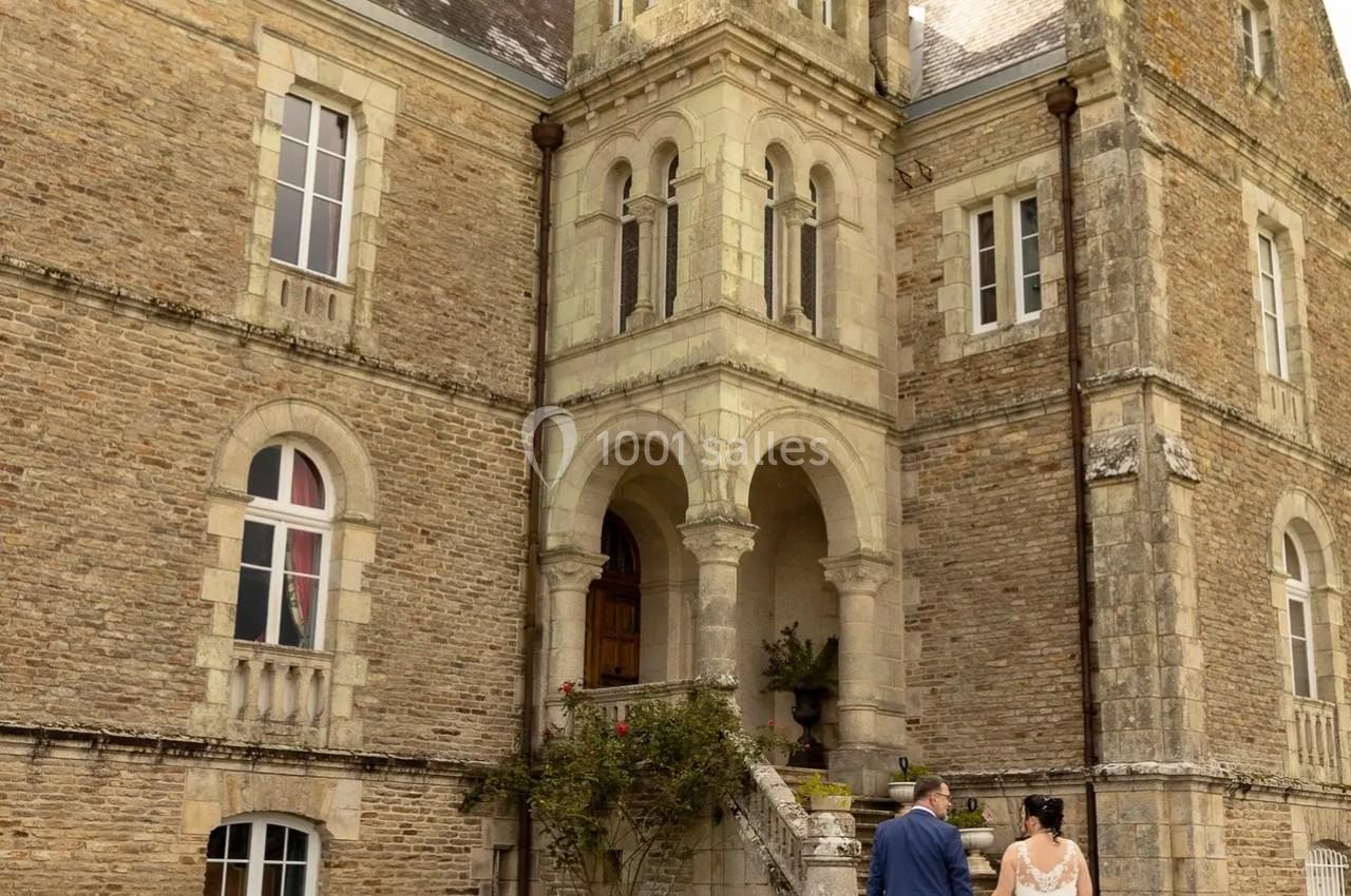 Un couple en tenue de mariage marche vers un château en pierre sous un ciel clair.