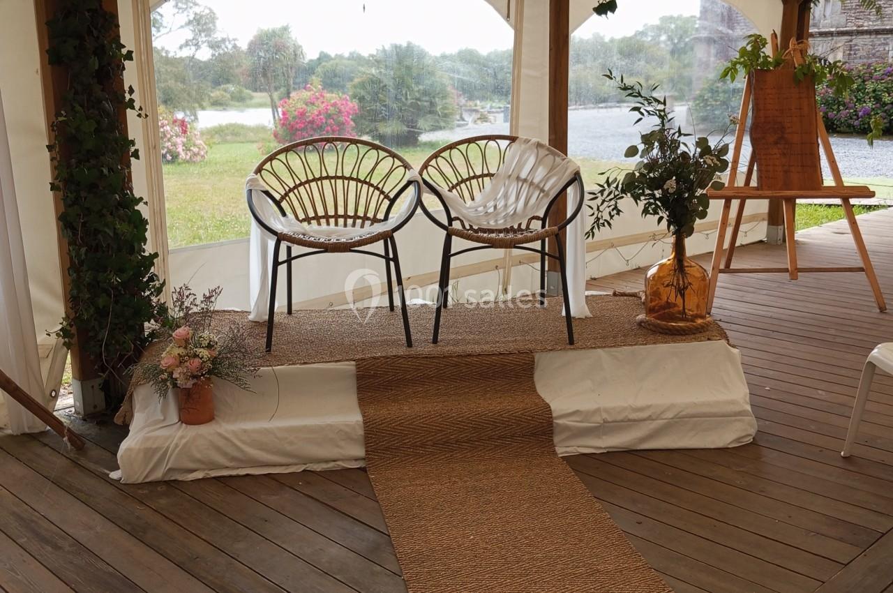 Deux chaises en rotin sur une estrade décorée de plantes, sous une tente ouverte avec vue sur un jardin et une rivière.