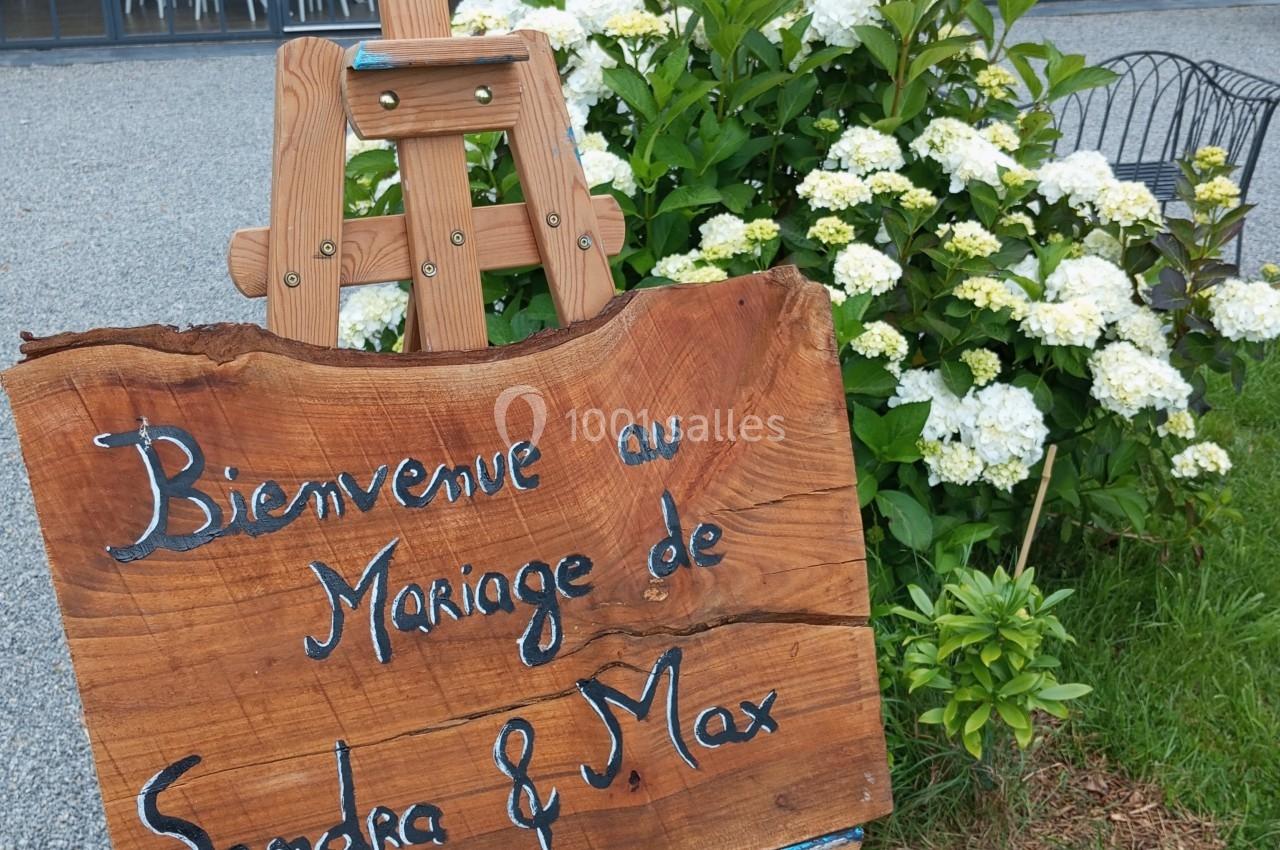 Panneau en bois sur chevalet indiquant ’Bienvenue au mariage de Sandra & Max’, devant un jardin et une salle vitrée.