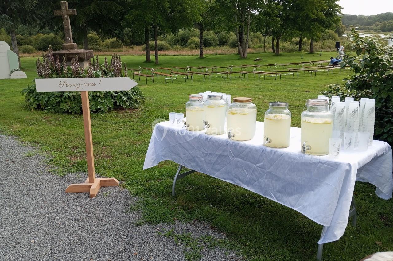 Table avec des bocaux de limonade et des verres, installée en extérieur près d'une flèche indiquant ’Servez-vous’.