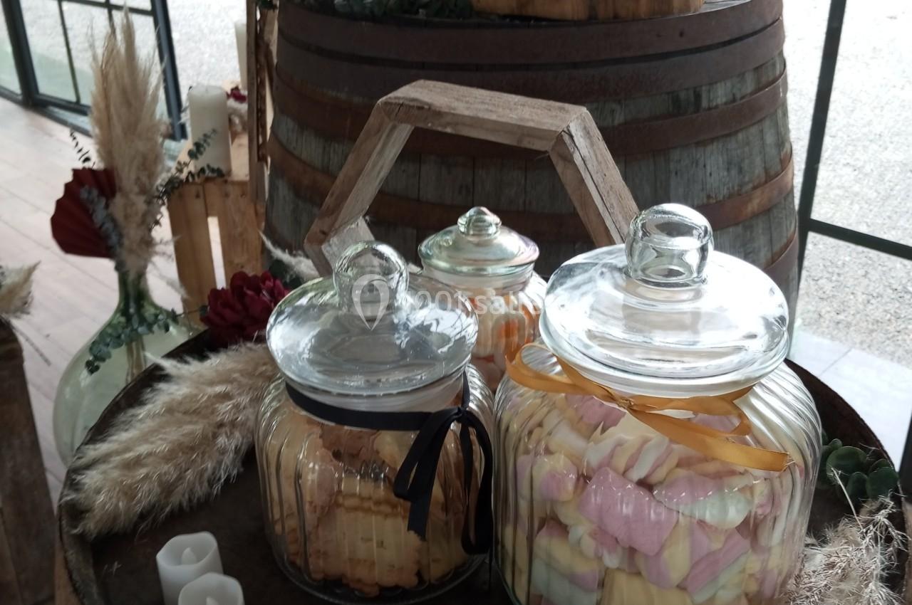 Bonbonnières en verre remplies de friandises colorées, posées sur des tonneaux en bois dans un décor champêtre.