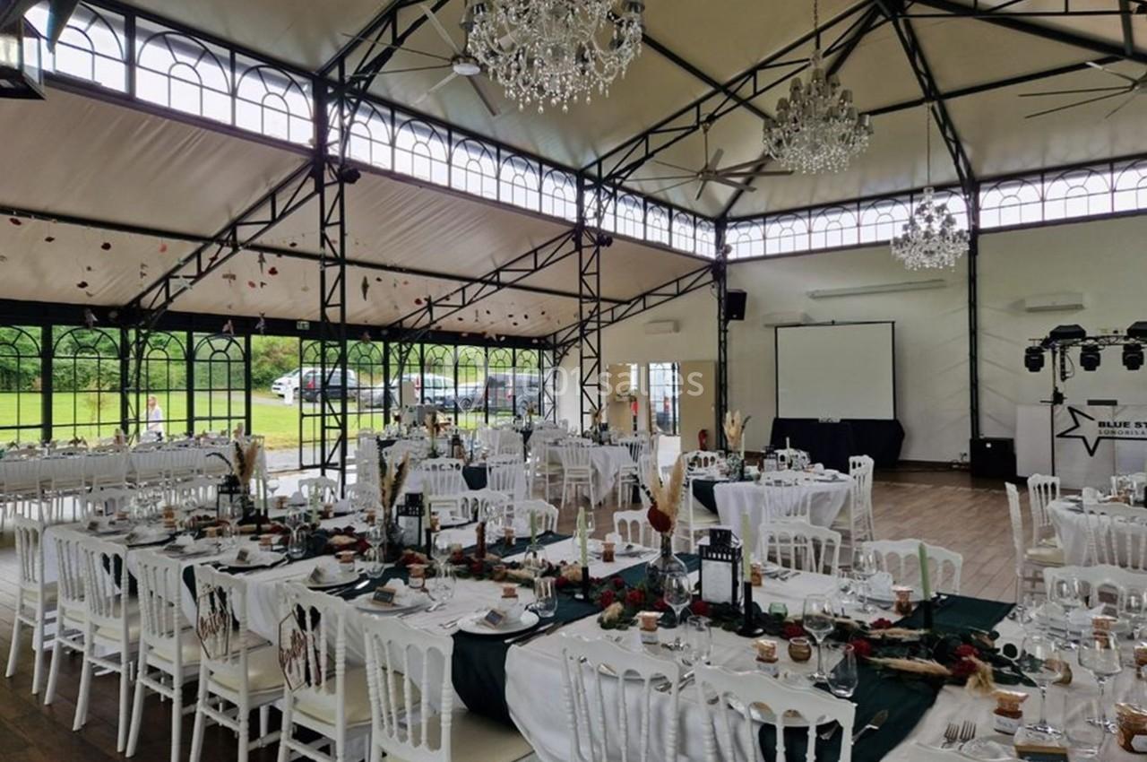 Salle de réception décorée pour un événement, avec tables dressées, chaises blanches et lustres suspendus.