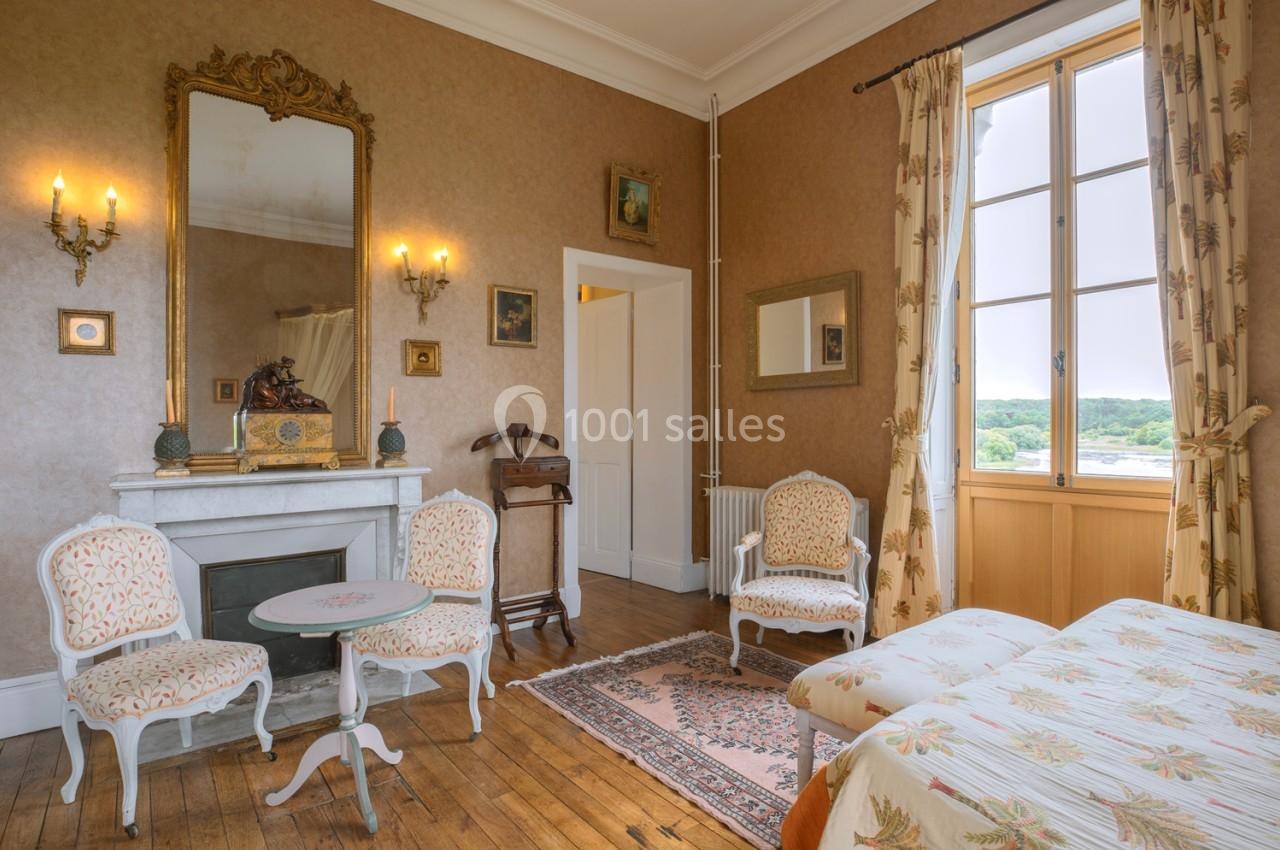 Chambre lumineuse avec mobilier ancien, cheminée, miroir doré, table et fauteuils, vue sur un paysage extérieur.