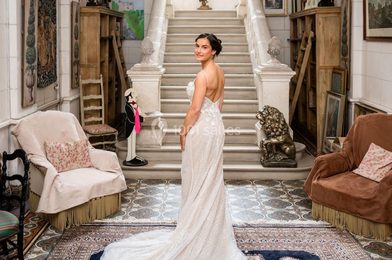 Femme en robe de mariée dans une pièce élégante avec tapis, fauteuils et escalier menant à une fenêtre lumineuse.