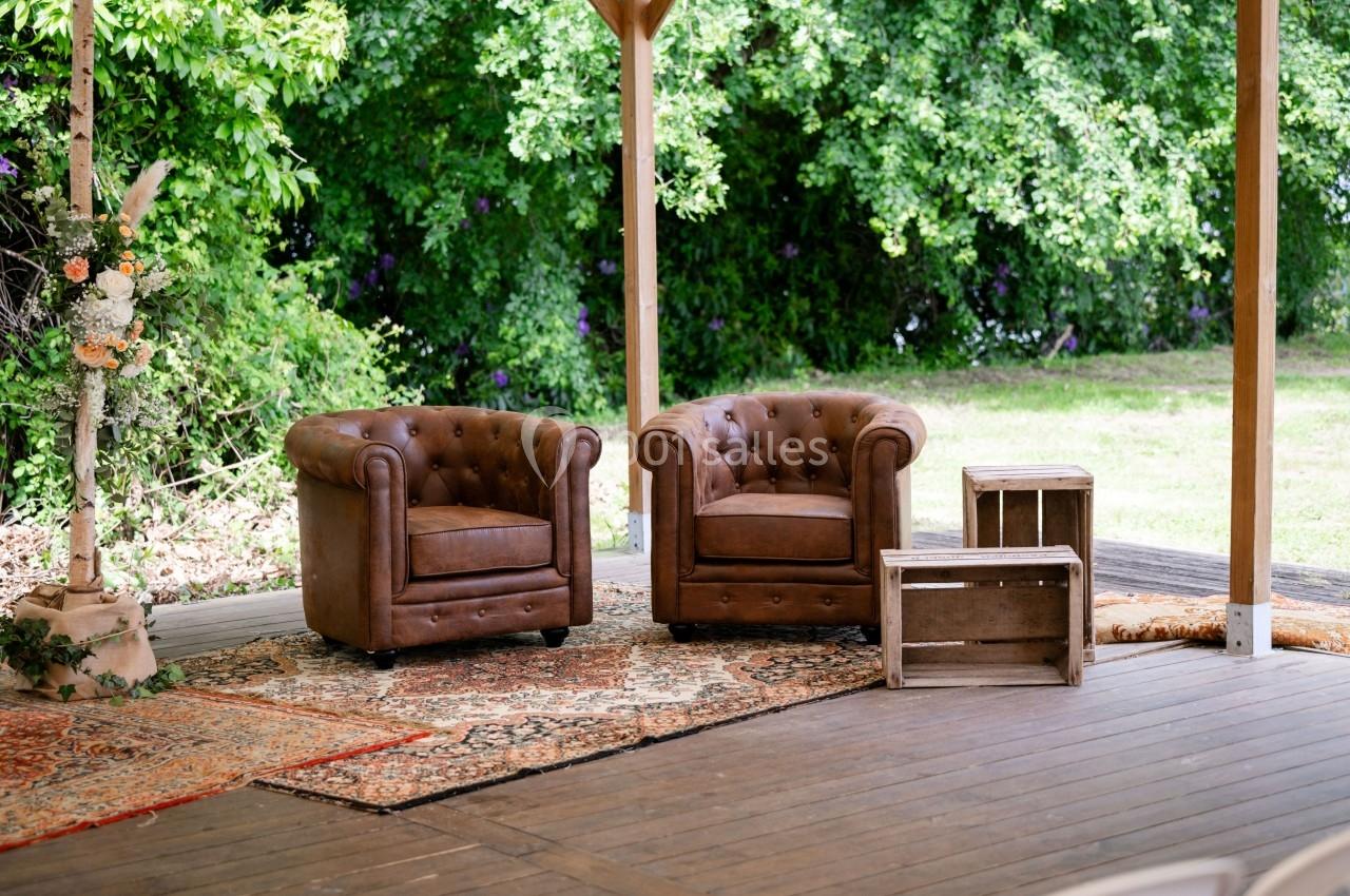 Deux fauteuils en cuir marron disposés sur un tapis, avec des caisses en bois, sous une structure en bois près d'arbres.