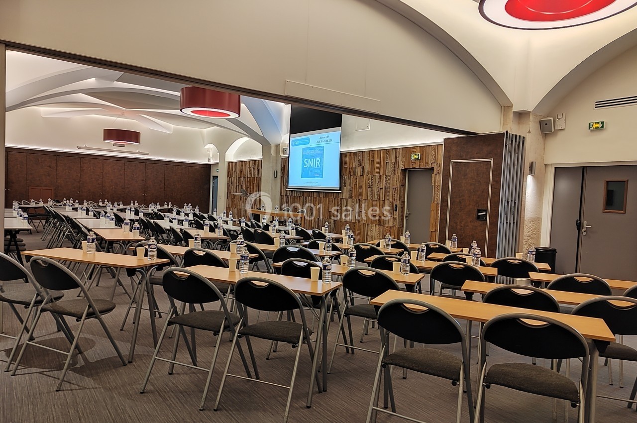 Salle de conférence avec des rangées de tables et chaises alignées, écran de projection allumé en arrière-plan.
