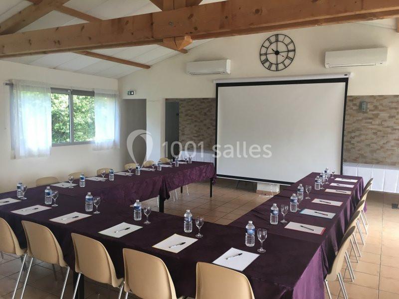 Salle de réunion lumineuse avec tables disposées en U, chaises, bouteilles d'eau, blocs-notes et écran de projection.
