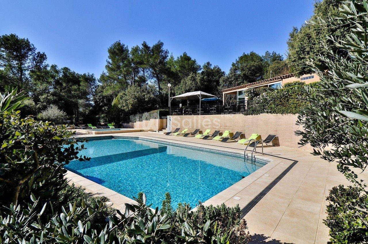 Piscine extérieure entourée de chaises longues, végétation et maison en arrière-plan sous un ciel dégagé.