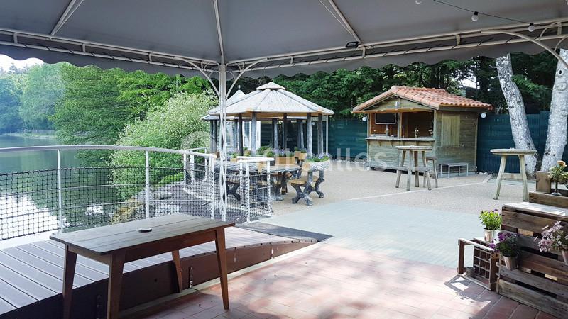 Terrasse couverte donnant sur un lac, avec des tables en bois et des kiosques en arrière-plan près d'une forêt.