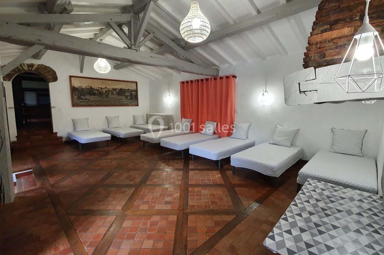 Salle avec des lits simples alignés, murs blancs, poutres apparentes, sol en carreaux de terre cuite et rideaux rouges.